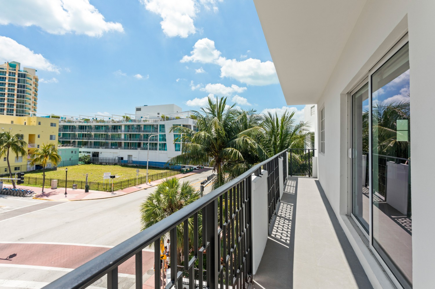A balcony with a black railing overlooks a street with palm trees and buildings in the background.