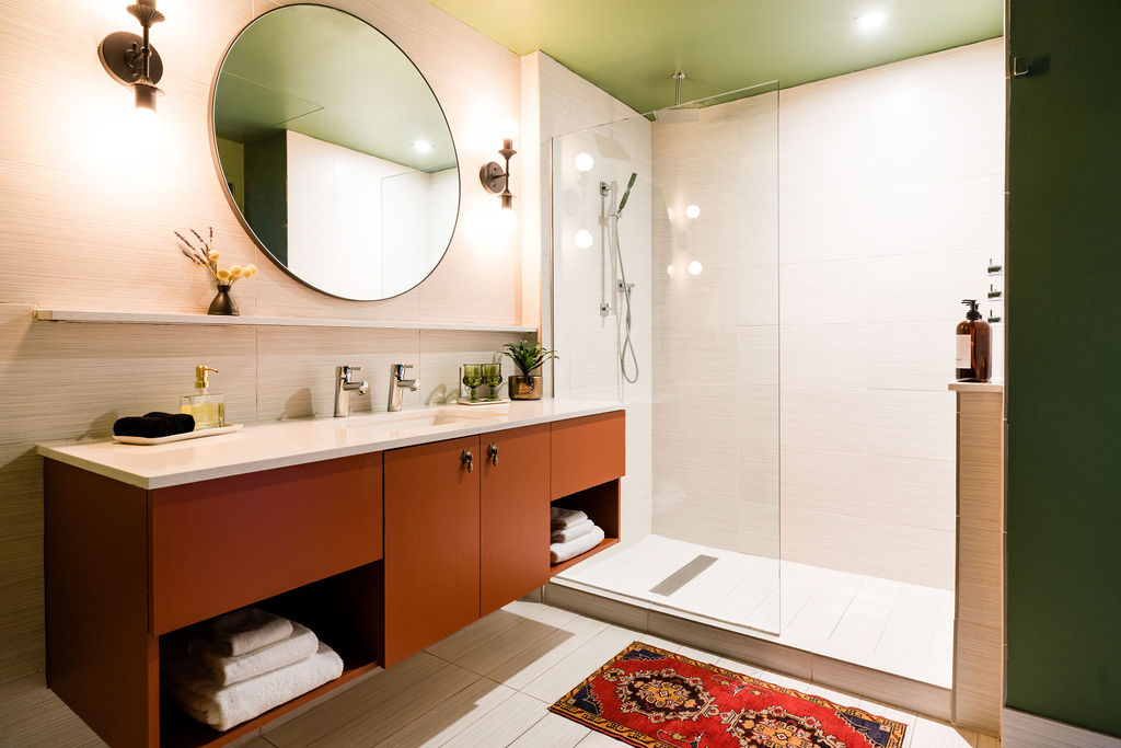 A modern bathroom with a double vanity, large round mirror, glass-enclosed shower, and colorful rug.
