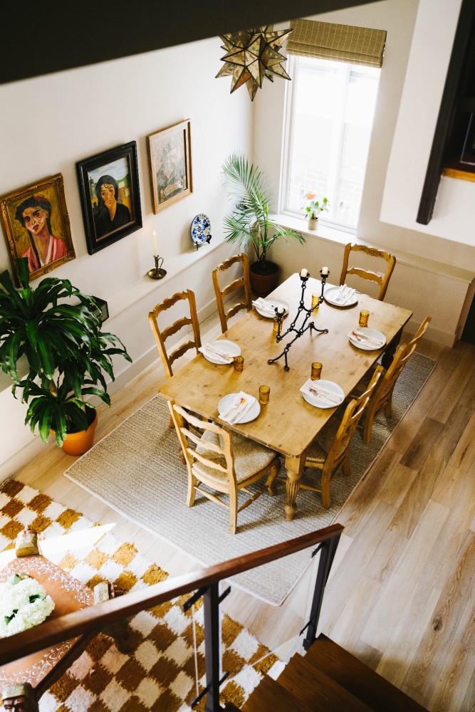 A dining area with a wooden table set for dinner, surrounded by chairs, potted plants, and artwork on the walls.