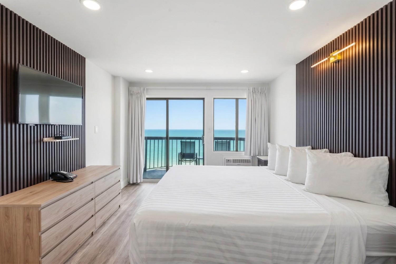 A wide-angle view of a hotel room with a king-sized bed, a large flat-screen TV mounted on the wall, a wooden dresser, and a balcony with ocean views.