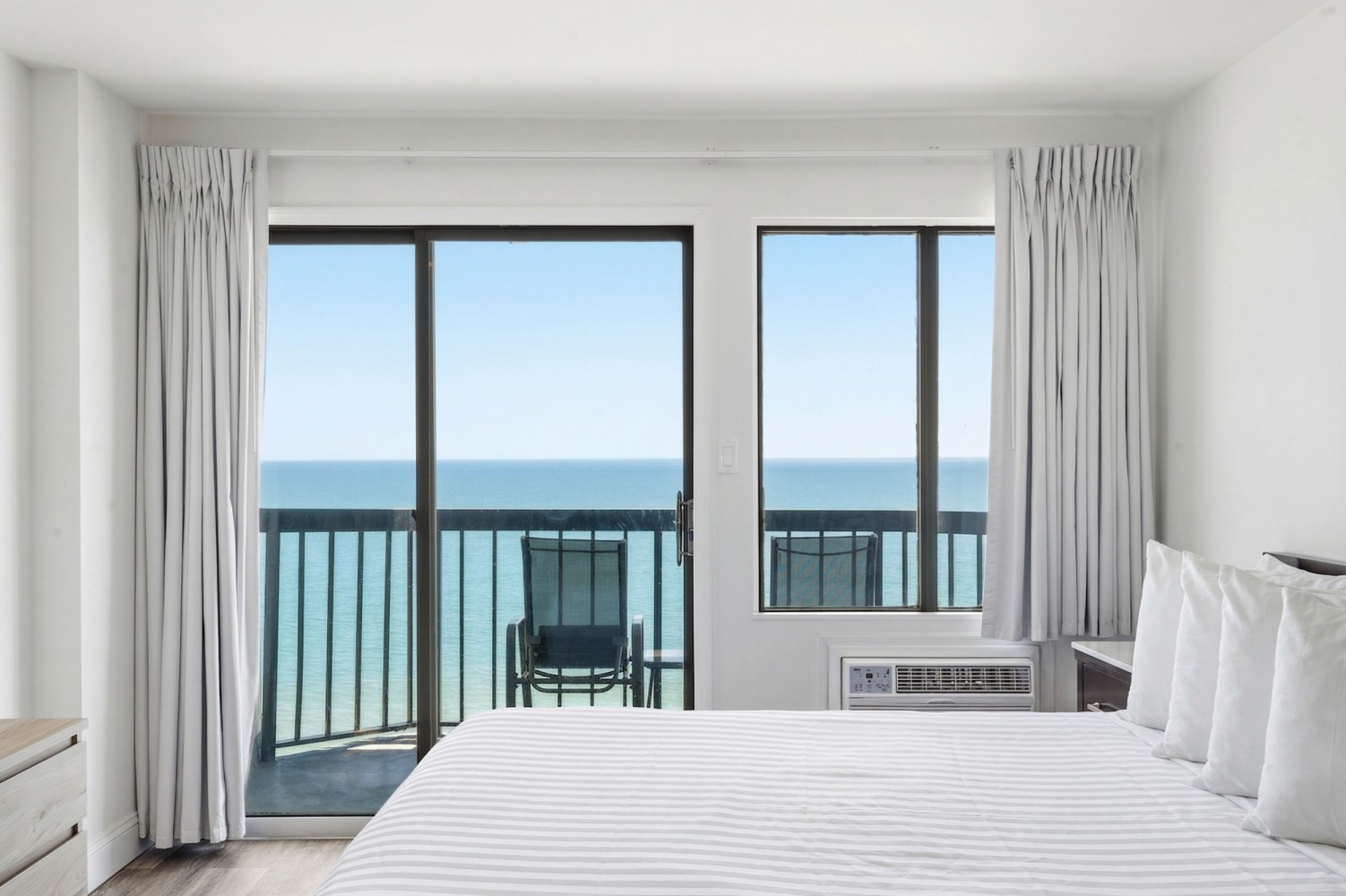 A wide view of a hotel room with a large bed, a window and sliding glass door leading to a balcony with an ocean view.