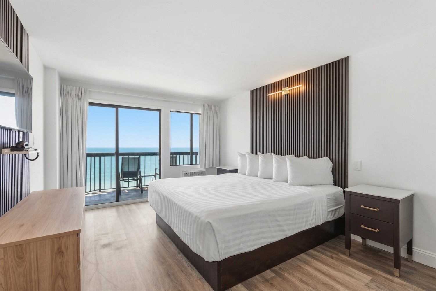 A modern hotel room with a king-sized bed, wooden floor, and a balcony overlooking the ocean.