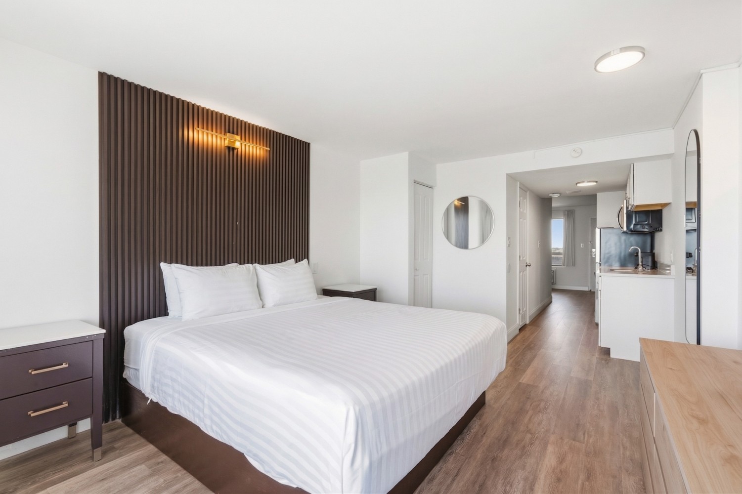 A wide view of a hotel room with a double bed, kitchenette, and wood floor.