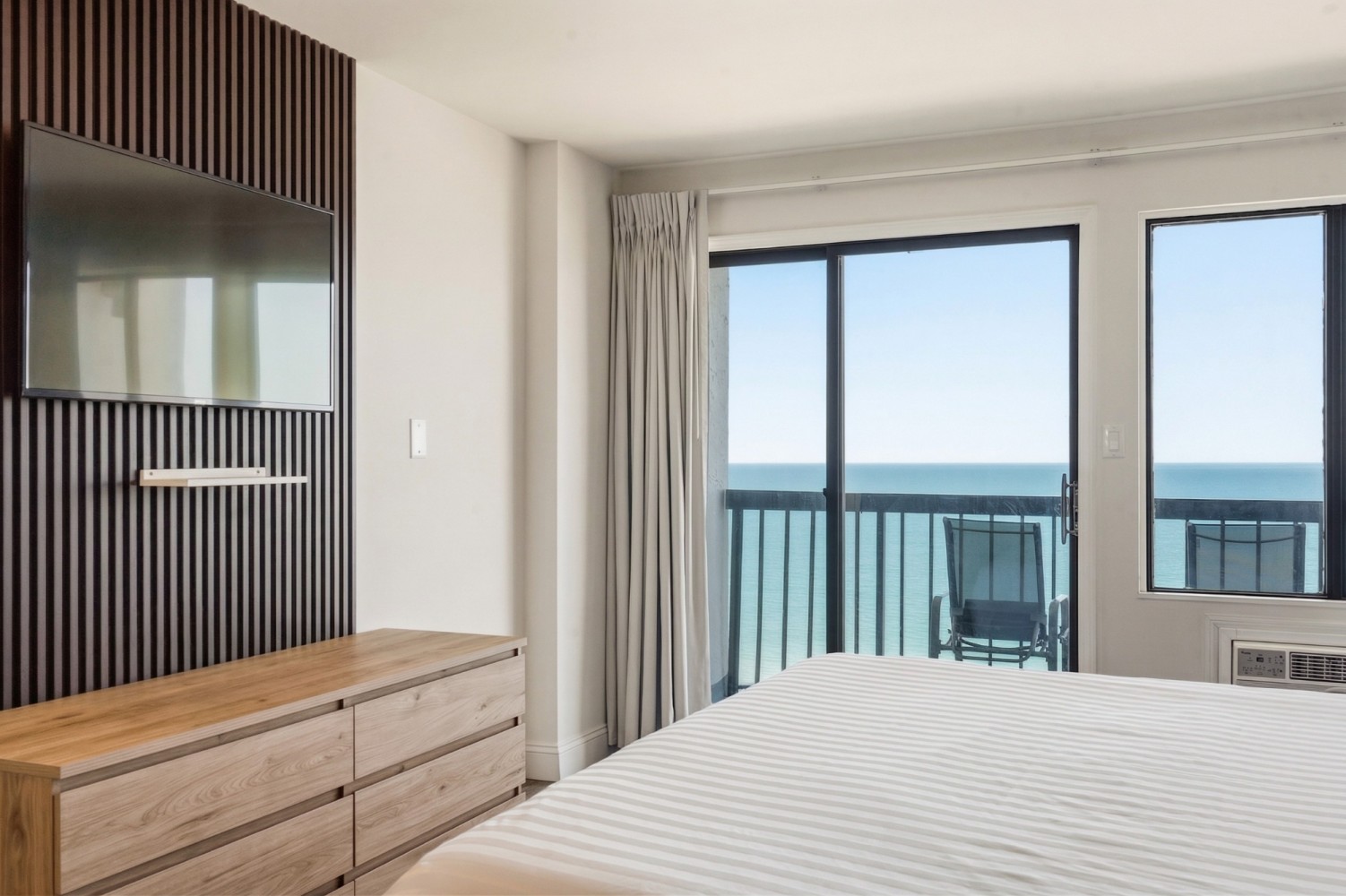 A hotel room with a large bed, wooden dresser, flat-screen TV, and sliding glass doors leading to a balcony with an ocean view.