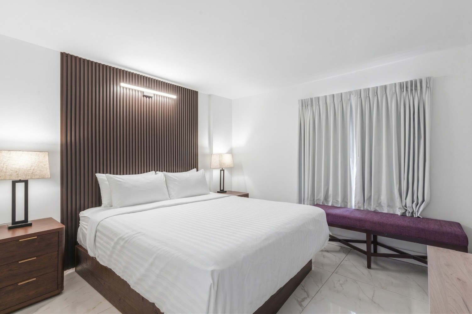 A hotel room with a large bed, two nightstands, a bench at the foot of the bed, and a window with curtains.