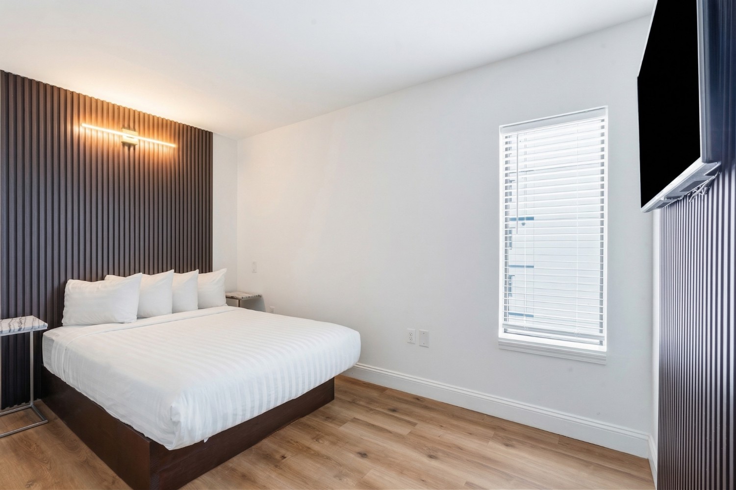 A wide-angle view of a hotel room with a bed, window, TV, and nightstand.