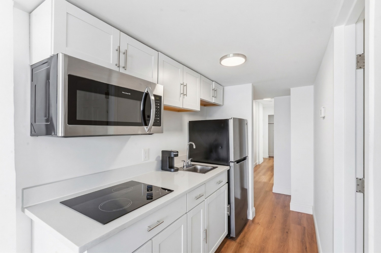 A narrow kitchenette with white cabinets, stainless steel appliances, and a black stovetop.