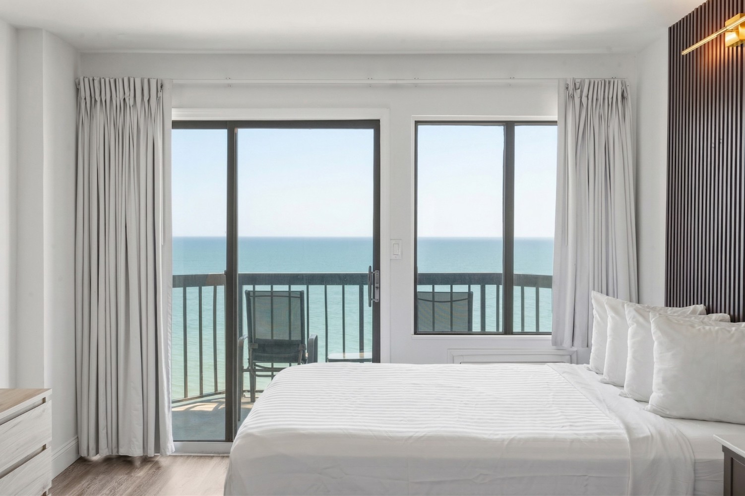 A wide view of a hotel room with a large bed, a sliding glass door leading to a balcony with an ocean view.