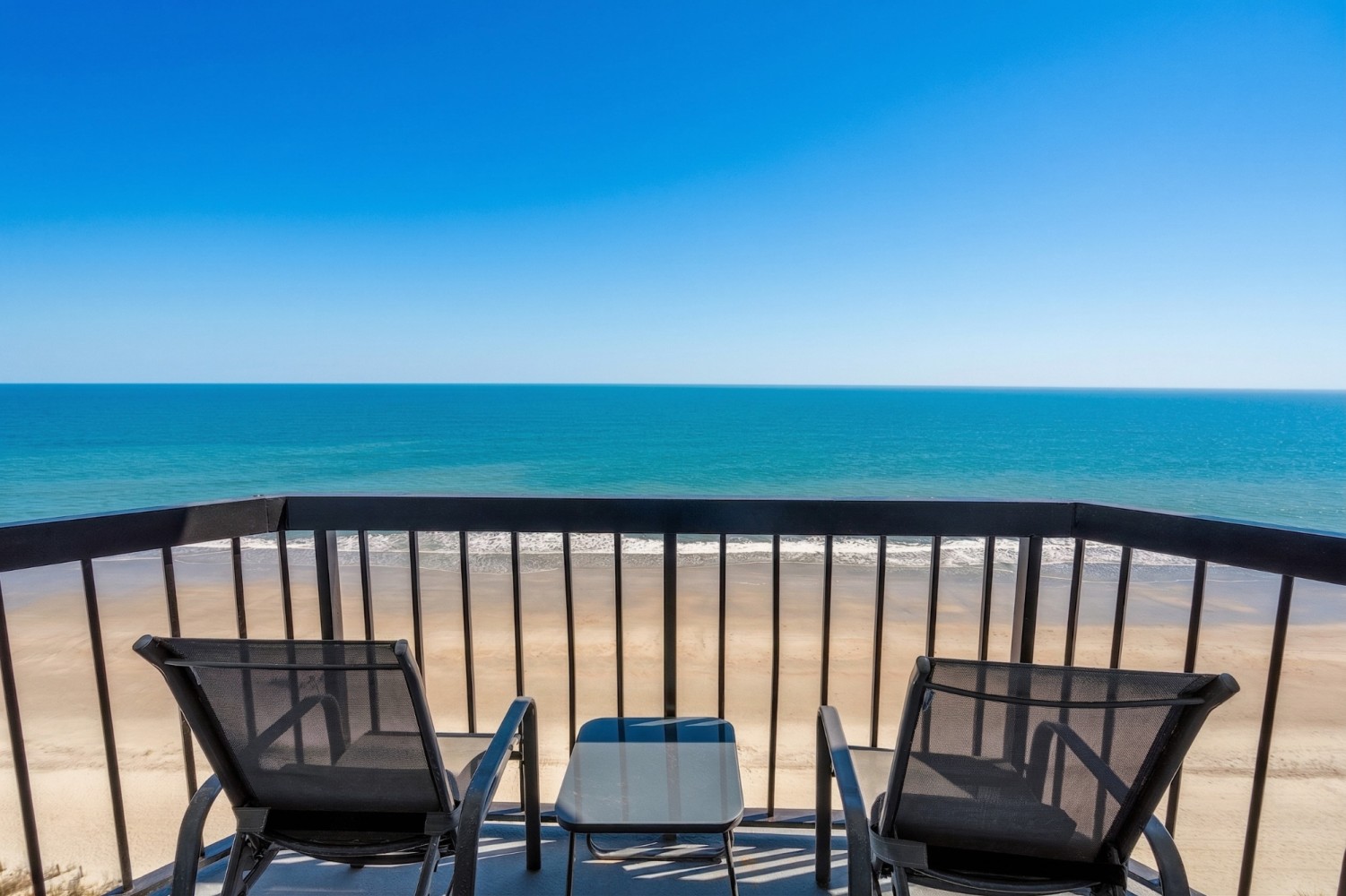 A balcony with two chairs and a small table overlooking a sandy beach and turquoise ocean.