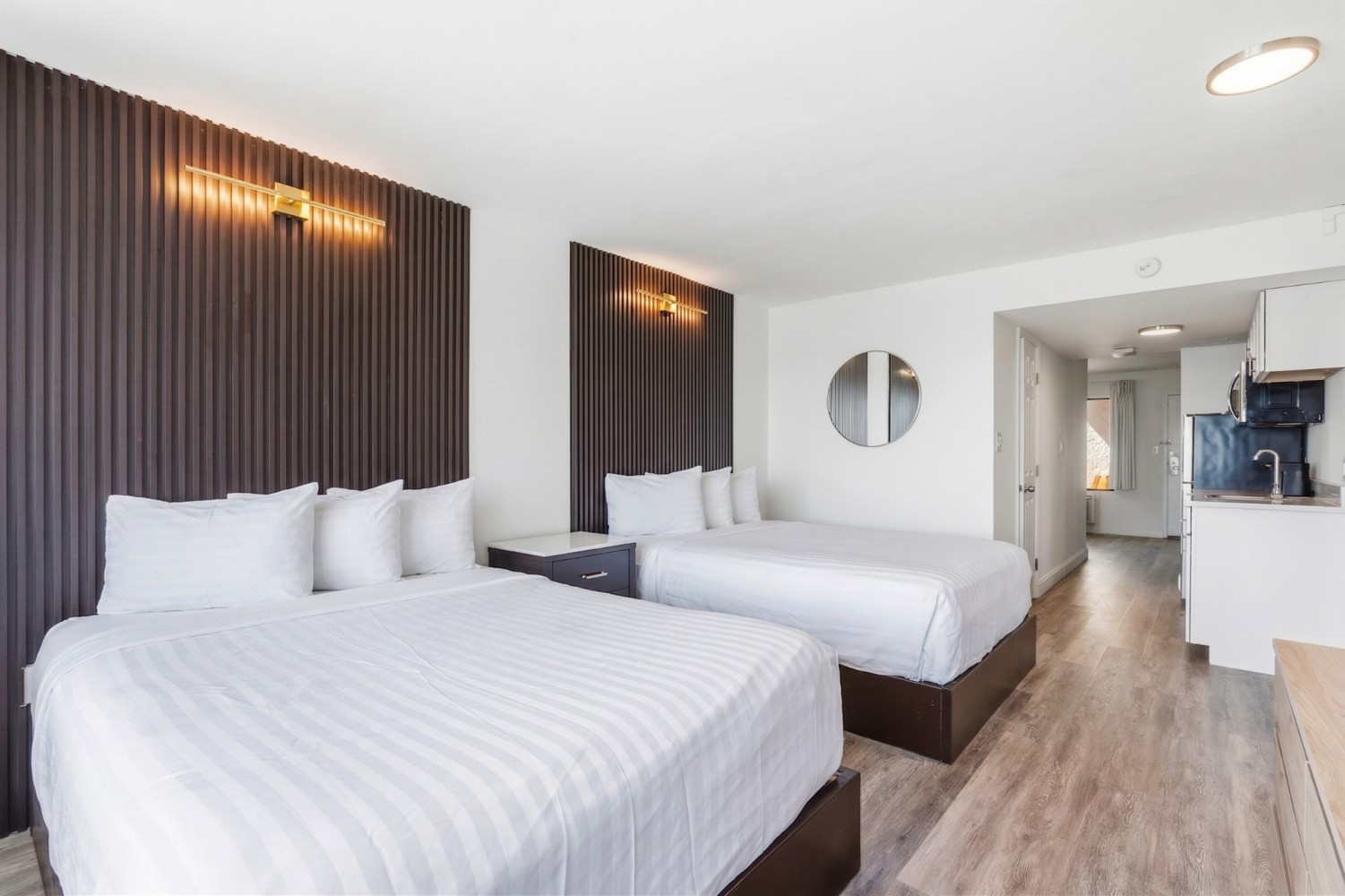A hotel room with two beds, a kitchenette, and modern lighting.