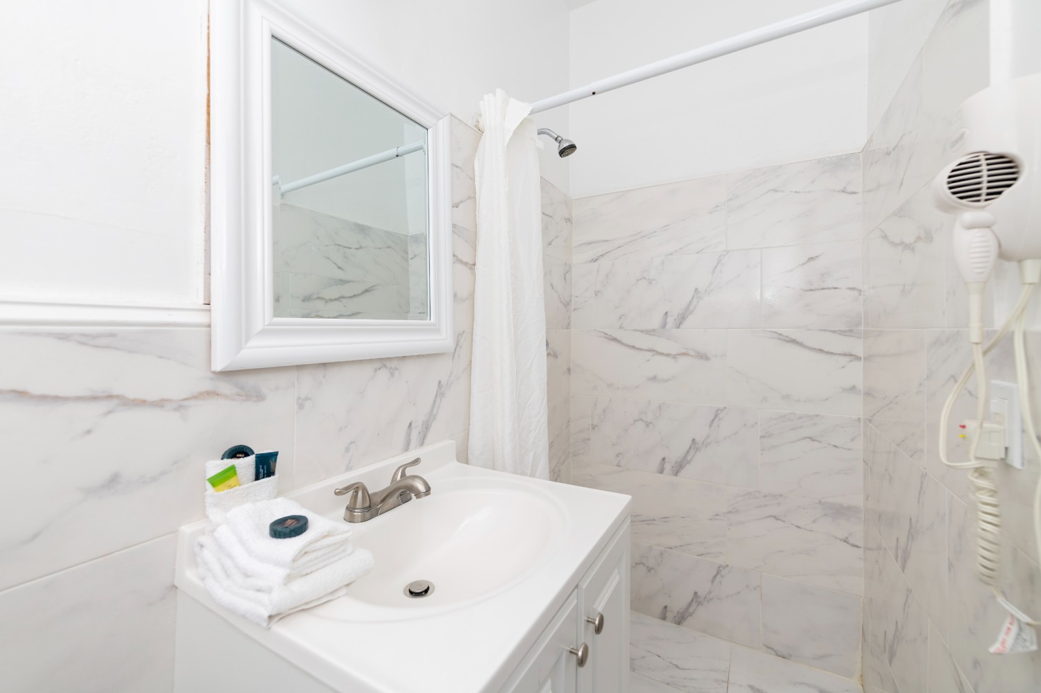 A clean and well-appointed bathroom with marble tiles, a shower with a white curtain, a sink with towels and toiletries, and a hair dryer.