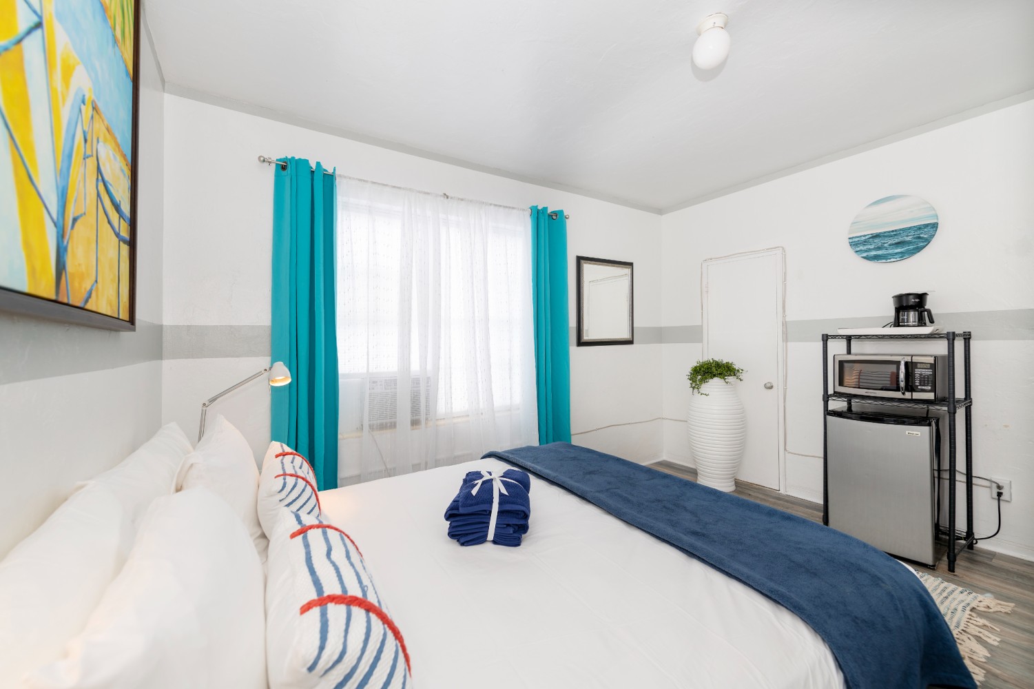 A hotel room with a king-size bed, a window with blue curtains, a coffee maker, and a small refrigerator.